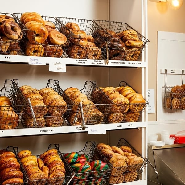 A display of various bagels in wire baskets on white shelves, with labels for each flavor.
