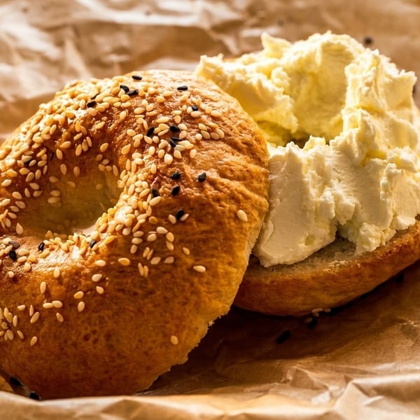 Sesame bagel with cream cheese on crumpled brown paper, illuminated by warm light.
