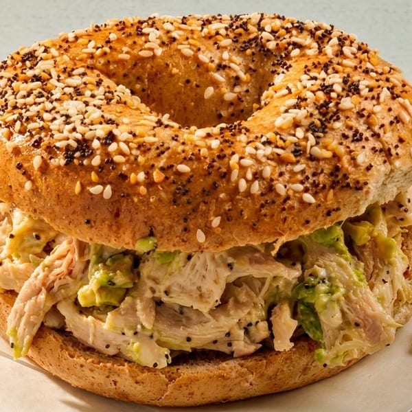 Everything bagel with chicken salad and avocado on parchment paper, on a light blue wooden surface.