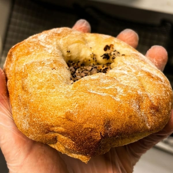A gloved hand holds a freshly baked, golden-brown everything bagel, with more bagels in the background.